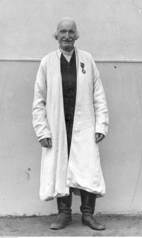 Jan Słomka, wójt z Dzikowa, 1929 rok. Na zdjęciu jest uśmiechający się mężczyzna w średnim wieku, w białej koszuli, czarnej kamizelce, wysokich, czarnych butach oraz długiej, białej, chłopskiej sukmanie, do której, z lewej strony piersi, przypięty jest medal. Mężczyzna ma wysokie czoło oraz siwe, odstające nad uszami włosy i siwe wąsy. Mężczyzna stoi przy budynku.