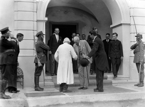 Prezydent Rzeczpospolitej Polskiej Ignacy Mościcki wita się z Janem Słomką, 1929 rok. Na zdjęciu jest grono elegancko ubranych osób na tle frontonu i wejścia budynku. Na pierwszym planie jest dwóch starszych, witających się w pokłonie prawymi dłońmi mężczyzn. Jeden z nich jest ubrany w elegancki, jasny garnitur, trzyma w lewej dłoni laskę, drugi - w długą, białą, chłopską sukmanę oraz wysokie, chłopskie, czarne buty. Tuż przy nich stoi w postawie „Baczność” żołnierz z karabinem zawieszonym na ramieniu. Nieco na prawo stoi drugi żołnierz, też w postawie „Baczność”, salutujący szablą ułożoną ku górze wzdłuż lewego obojczyka.