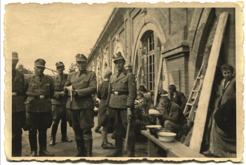 Na archiwalnej fotografii grupa Niemców w mundurach pozuje do zdjęcia z bronią w ręku, w tle pozbawieni wolności ludzie tłoczą się pod murami fortu.