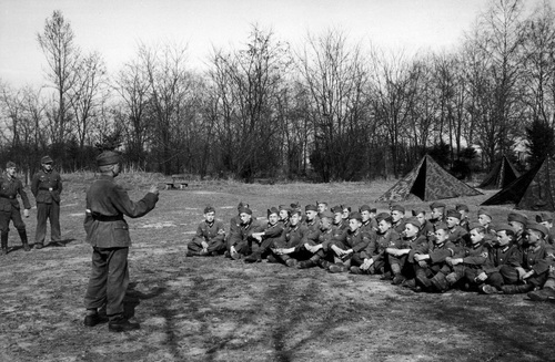 Obóz młodzieży z Hitlerjugend w Kobierzynie. Widoczna grupa siedzących na trawie chłopców w mundurkach. W tle rozbite namioty wojskowe. Fot. NAC