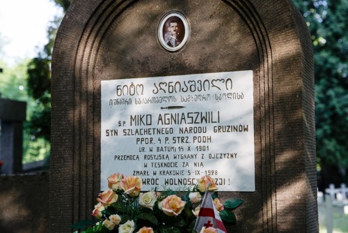 Współczesne zdjęcie z widocznym fragmentem nagrobka, na którym umieszczono napisy w języku polskim i gruzińskim oraz zdjęcie zmarłego. W dolnej części widoczne kwiaty z dekoracją w postaci niewielkich rozmiarów flagą Gruzji.