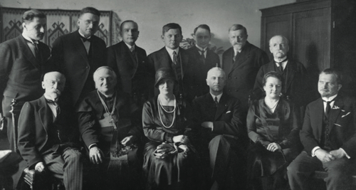 Grupa osób w dwóch rzędach, pozująca do zdjęcia. Osoby w pierwszym rzędzie siedzą, wsród nich dwie elegancko ubrane kobiety i mężczyzna w stroju hierarchy katolickiego