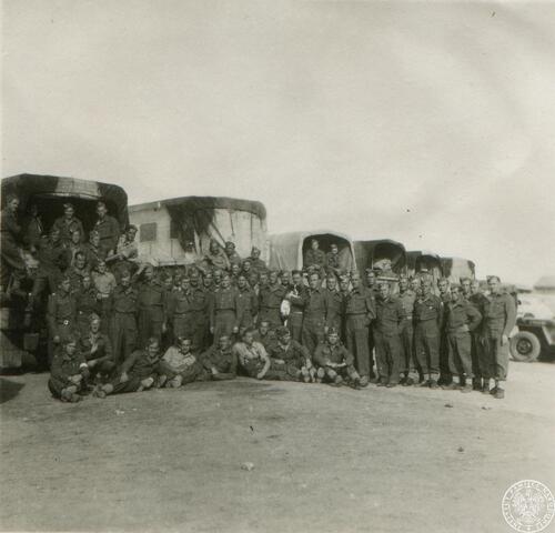 Grupa żołnierzy pozuje do zdjęcia w otwartej przestrzeni. Za ich plecami pojazdy wojskowe.
