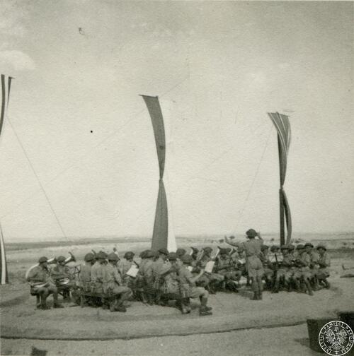 W otwartej przestrzeni orkiestra wojskowa gra prowadzona przez dyrygenta. Za orkiestrą długie, pionowo umieszczone, tkaniny pełniące funkcję flag, w tym jedna biało-czerwona.