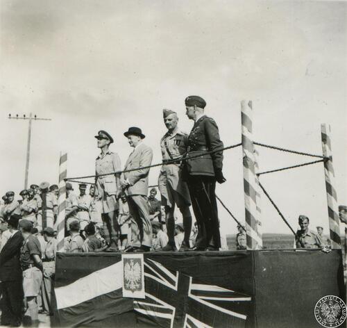 Na improwizowanej trybunie stoją czterej mężczyźni, w tym jeden w ubraniu cywilnym (garnitur i kapelusz) i trzech w mundurach oficerskich: dwóch polskich i jeden - brytyjskim. Wokół trybuny stoją żołnierze. Trybuna udekorowana jest flagami: polską i brytyjską oraz godłem polskim.