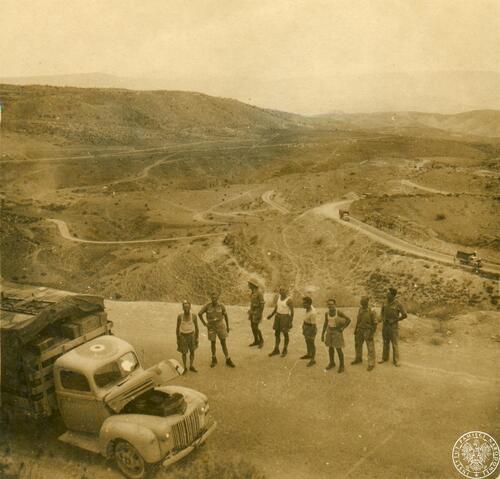 Grupa mężczyzn, wojskowych, stoi w otwartym terenie, na drodze, przed ciężarówką wojskową. W tle teren pagórkowaty, przez który przebiegają drogi.