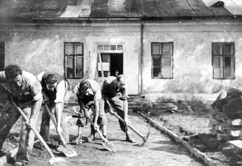 Czterech pochylonych mężczyzn wyrównuje teren łopatami i kilofami przed przeznaczonym do rozbiórki parterowym domem. Mężczyźni ubrani są w normalne ubrania: spodnie i koszule.