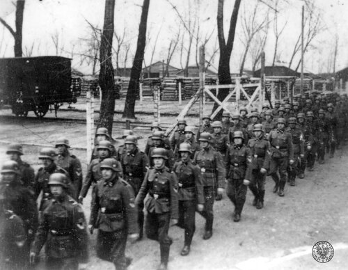 Na pierwszym planie kolumna niemieckich żołnierzy SS przechodzi wzdłuż ogrodzenia z drutu kolczastego. Nieco w głębi stojący na torach wagon. Dalej, w tle, drewniane budynku, a przed nimi widać pryzmy z drewnem.