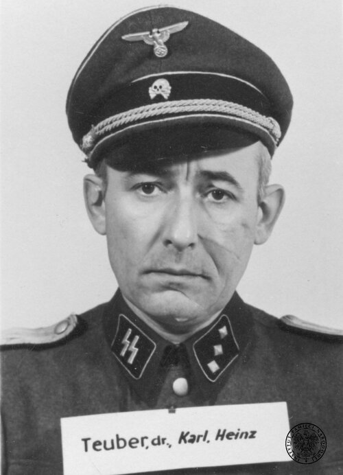 Zdjęcie portretowe gładko ogolonego, z długimi bliznami na twarzy mężczyzny w mundurze oficera niemieckiego ze znakiem „SS” na prawym kołnierzu munduru oraz w czapce z trupią główką i niemieckim nazistowskim orłem. Na wysokości piersi tabliczka ze słowami: „Teuber, dr., Karl, Heinz”.