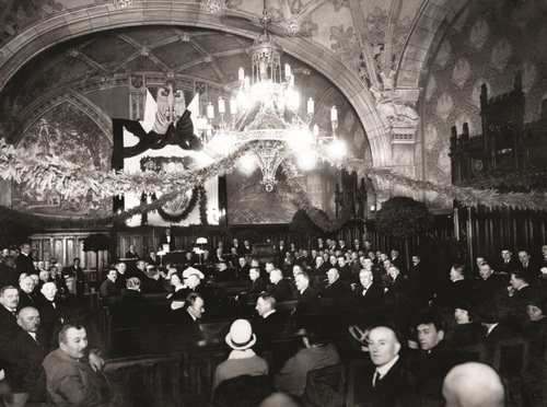 Uroczyste, 99. posiedzenie I Sejmu Śląskiego w Katowicach, 10 października 1924 r. Posłowie zasiadający w ławach na sali obrad. W stylowym pomieszczeniu wyróżnia się okazały żyrandol. Na ścianie za prezydium wizerunek Orła.