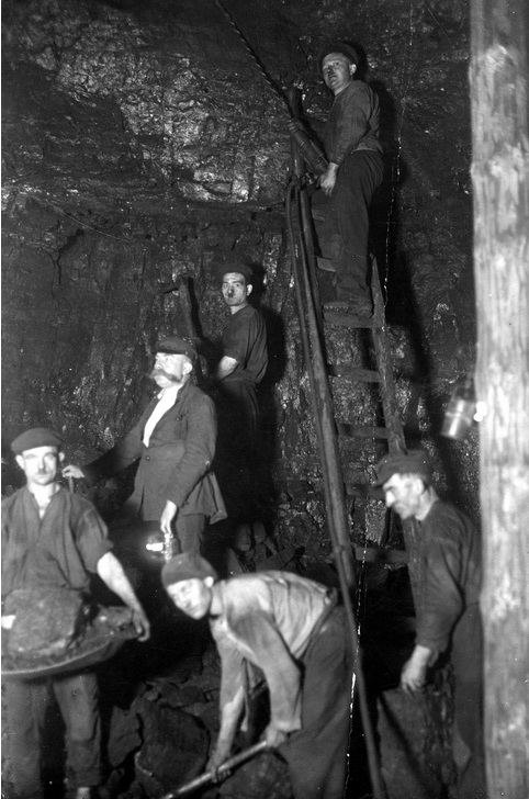 Górnicy przy pracy na ścianie kopalnianego tunelu. W rękach trzymają narzędzia służące do skuwania węgla.