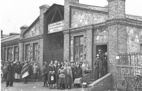 Rodziny strajkujących oczekują przed bramą kopalni węgla kamiennego &quot;Klimontów&quot; w Sosnowcu, 1933 r. Kobiety w typowych strojach stoją przed ceglanym budynkiem okalającym bramę. Na schodach wejścia do budynku funkcjonariusze policji.