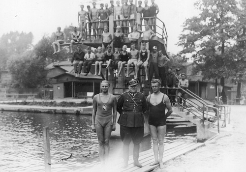 Przedolimpijski kurs pływacki w Tychach, lipiec 1934 r. Fotografia grupowa uczestników i kierownictwa przedolimpijskiego kursu pływackiego. Na pierwszym planie trójka osób nad krawędzią zbiornika wodnego. W głębi grupa osób zajmujących miejsca na krawędziach wieży służącej do wykonywania skoków.