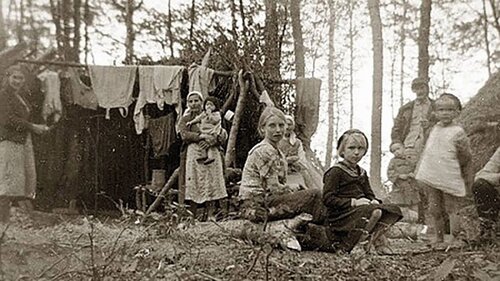 Trzy kobiety, mężczyzna, dzieci; jedna z kobiet trzyma na rękach małe dziecko. Rodzina? W lesie improwizowany namiot, na którym suszą się ubrania