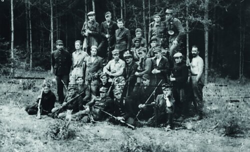 Grupa mężczyzn, z których tylko parę osób nosi ubrania cywilne, a zdecydowana większość - wojskowe; większość trzyma w rękach broń. Zdjęcie na tle lasu