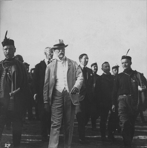 Prezydent w jasnym garniturze kroczący wśród grupy mężczyzn w charakterystycznych strojach organizacji sokolskiej.