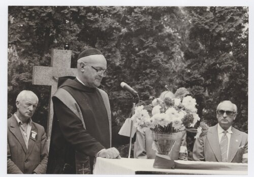 Ksiądz/zakonnik przemawia przy ołtarzu przystrojonym bukietem kwiatów. Z tyłu stoi dwóch widocznych mężczyzn i jedna osoba niewidoczna. Za księdzem/zakonnikiem - kamienny krzyż