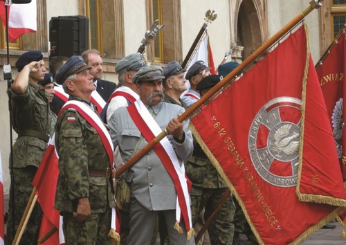 Grupa osób, przepasanych biało-czerwonymi szarfami, przy budynku. Kilka z nich nosi rekonstrukcyjne mundury legionowe i legionowe czapki. Widać dwa, pochylone, sztandary; w tle są dwa kolejne, widoczne tylko w mniejszych fragmentach