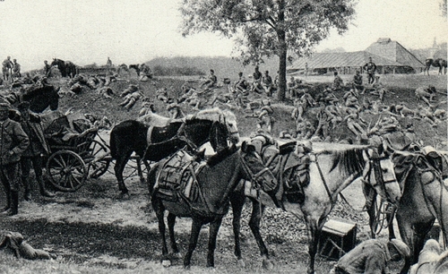 Odpoczynek oddziałów legionowych pod Jastkowem, 1915 rok. Pagórkowate pole, na którym bezpośrednio na ziemi leżą i odpoczywają żołnierze. Wśród nich stoją osiodłane konie wojskowe. Widać również zaprzęg konny ze stojącymi obok paroma żołnierzami. W tle zabudowania.