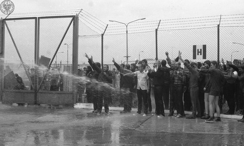 Akcja protestacyjna internowanych w ośrodku odosobnienia w Kwidzynie w dniu 14 sierpnia 1982 r. Polewani są wodą prawdopodobnie z węża strażackiego. Część z nich ma uniesione ręce z palcami w geście "V". Na drugim planie, wysoki płot za nim  także stoją mężczyźni. Z lewej strony zamknięta brama. Fot. AIPN