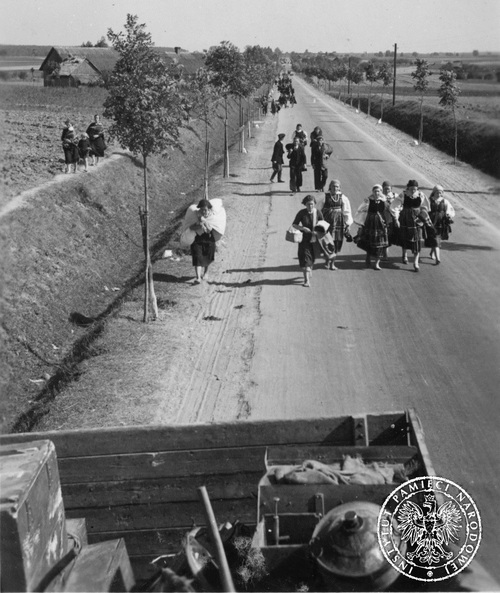 Wrzesień 1939 roku, okolice Łaznowa. Przemieszczający się pieszo, objuczeni tobołami, polscy uchodźcy wojenni na asfaltowej drodze, na trasie marszu oddziałów niemieckich. Niektóre z idących kobiet ubrane są w tradycyjne stroje ludowe. W tle zabudowania.
