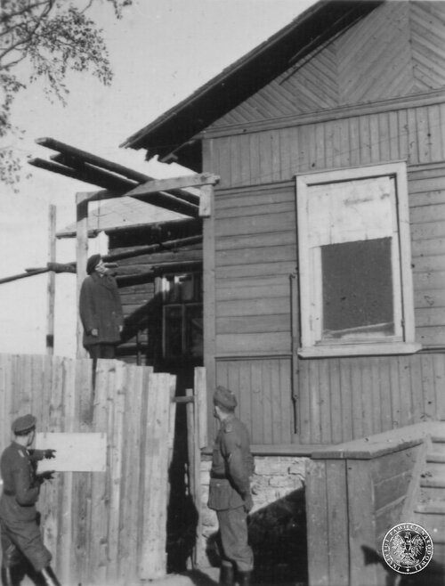 Powiat lipnowski lub sierpecki, 1939 r. Mężczyzna powieszony na szubienicy obok drewnianego domu, na pierwszym planie dwóch żołnierzy niemieckich. Fot. z zasobu IPN