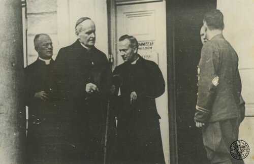 Biskup pomocniczy warszawski i wikariusz kapitulny archidiecezji warszawskiej Antoni Szlagowski wizytuje obóz Dulag 121 w Pruszkowie, 20 sierpnia 1944 roku. Biskup ma piuskę na głowie, ubrany jest w płaszcz, spod którego widać koloratkę; w rękach trzyma laskę. Przy nim stoi dwóch innych kapłanów, także ubranych w płaszcze, z widocznymi spod nich koloratkami. Z boku stoją jeszcze dwaj inni mężczyźni, w tym jeden to być może jakiś niemiecki funkcjonariusz. W tle fragment budynku i drzwi wejściowe.