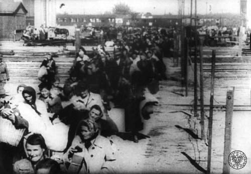 Wejście do obozu w Pruszkowie wypędzonych z Woli. Mieszkańcy Woli, objuczeni bagażami z resztkami dobytku, przechodzą torami kolejowymi. Wśród idących dominują kobiety. W głębi wagony pociągu.