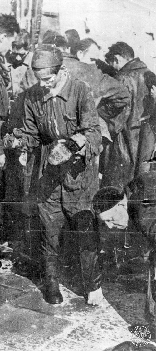 Polacy - więźniowie Niemców w „Durchgangslager 121 Pruszków”. Nędznie ubrani ludzi stoją na placu. Jedna z postaci jest pół bosa, trzymając w rękach i czyszcząc lub naprawiając but.