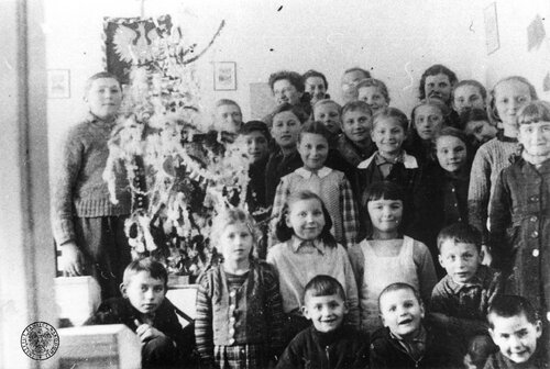 A group of Polish children and adults (teachers?) at the public school for refugees at Kadarkut (Hungary) – commemorative picture next to a Christmas tree, December 1940. In the background Poland’s emblem hangs on the wall. The photo print gifted by Wanda Cichoszewska from Nowy Sącz, during the war a teacher at the school. Photo from the collection of the Institute of National Remembrance