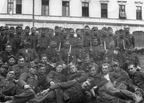 Żołnierze polscy i węgierscy pozują do grupowego zdjęcia na tle piętrowego budynku. Część posiada broń z nałożonymi bagnetami. Polacy są prawdopodobnie jeńcami wojennymi w obozie na Węgrzech. Fot. z zasobu IPN (dar prywatny Marii Florczyk)