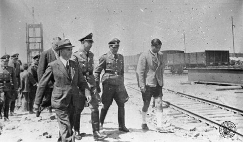 Szef SS Heinrich Himmler (3P) i komendant obozu w Oświęcimiu major SS (SS-Sturmbannführer) Rudolf Höss (2P) z innymi oficerami SS i przedstawicielami kierownictwa budowy (w tym mężczyzna w krótkich spodniach i białych podkolanówkach) przechodzą przez teren dworca kolejowego budowy zakładów Buny. Na pierwszym planie w kapeluszu szef budowy doktor Max Faust. Pierwszy z lewej wyższy dowódca SS i policji we Wrocławiu, generał broni SS Ernst Schmauser. Z tyłu widać grupę więźniów. Fot. z zasobu IPN
