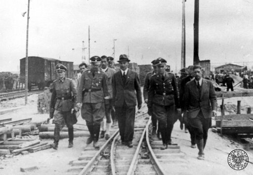Szef SS Heinrich Himmler (2L z przodu) i kierownik budowy doktor Max Faust (z przodu w kapeluszu) opuszczają teren budowy I.G. Farbenindustrie w Oświęcimiu („Buna-Werke”) idąc wzdłuż toru kolejki wąskotorowej. Za nimi przedstawiciele kierownictwa budowy i oficerowie SS, w tym komendant obozu w Oświęcimiu SS-Sturmbannführer Rudolf Höss (z przodu 2P) i oficer personalny szefa SS SS-Obersturmbannführer Rudolf Brandt (3 za Himmlerem). W tle drewniane budynki i pracujący ludzie. Fot. z zasobu IPN