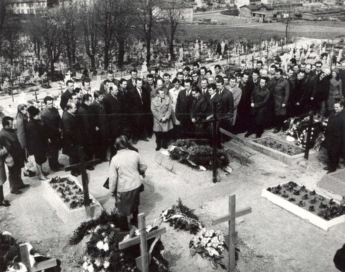 Delegacja gdańskich stoczniowców składa wieńce na grobie zabitych w Grudniu 1970 na cmentarzu świętego Franciszka w Gdańsku-Emaus, 8 maja 1971 roku. Na zdjęciu widać fragment cmentarza ze świeżymi, bez nagrobków, ale z krzyżami, grobami, na których widać ślady ich pielęgnowania. Przy grobach tych stoi bardzo liczne grono osób ubranych odświętnie, w koszule, krawaty, płaszcze. W tle zabudowa, nieco bliżej, już w obrębie cmentarza, drzewa.
