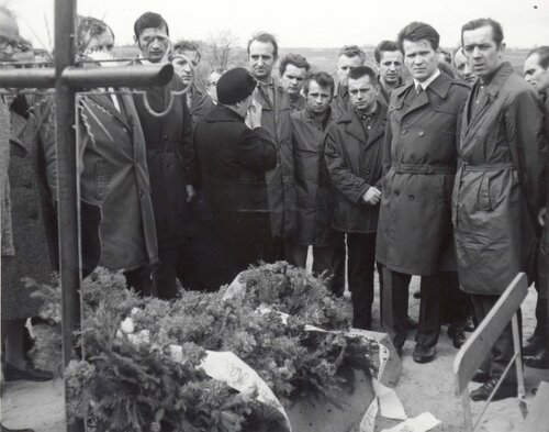 Delegacja gdańskich stoczniowców składa wieńce na grobie zabitych w Grudniu 1970 na cmentarzu św. Franciszka w Gdańsku-Emaus, 8 maja 1971 roku. Na zdjęciu widać grupę mężczyzn, ubranych w płaszcze, stojących w powadze nad świeżym, pokrytym wieńcami grobem bez nagrobka, ale z metalowym krzyżem. Plecami do fotografującego stoi ubrana na czarno kobieta wycierająca chusteczką oczy.