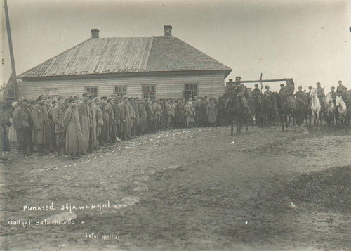 Jeńcy wojenni przed Stanisławem Bułakiem-Bałachowiczem, październik-listopad 1919 roku. Rzędy stojących jeńców. Przed nimi, na koniach, żołnierze Stanisława Bułaka-Bałachowicza, w tym on sam. Z tyłu drewniany, duży budynek.