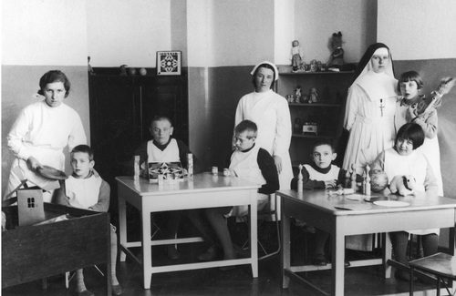 Dzieci i personel szpitala psychiatrycznego w Chełmie, 1936 rok. Na zdjęciu widać salkę edukacyjną ze stolikami, przy których siedzą, bawiąc się i ucząc zabawkami, dzieci - dziewczynki i chłopcy - w fartuszkach. Obok dzieci stoją trzy kobiety: dwie pielęgniarki, z których jedna ma czepek na głowie, oraz siostra zakonna w habicie z widocznym, zawieszonym na szyi, krzyżem. W głębi półka z zabawkami. Fotografia ze zbiorów Narodowego Archiwum Cyfrowego.
