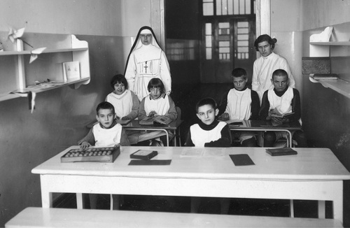 Dzieci i personel szpitala psychiatrycznego w Chełmie, 1936 rok. Na zdjęciu widać salkę edukacyjną ze stołami, przy których siedzą, ucząc się z książek i liczydła, dzieci - dziewczynki i chłopcy - w fartuszkach. Obok dzieci stoją dwie kobiety: pielęgniarka oraz siostra zakonna w habicie z widocznym, zawieszonym na szyi, krzyżem. Po obydwu stronach salki, na ścianach, wiszą półki. W głębi - drzwi wejściowe. Fotografia ze zbiorów Narodowego Archiwum Cyfrowego.
