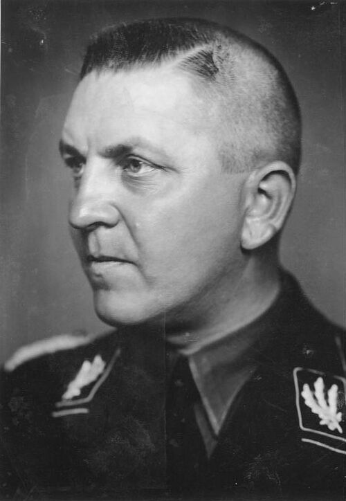 Theodor Eicke (1892-1943). Na zdjęciu jest młody mężczyzna, gładko ogolony i o krótko ostrzyżonych włosach, ubrany w mundur policyjnej formacji niemieckiej z okresu Niemiec hitlerowskich.
