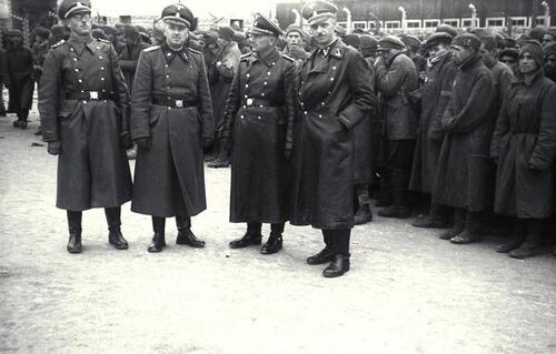 SS-Totenkopfverbände. Na zdjęciu widać czterech oficerów formacji niemieckich z okresu Niemiec hitlerowskich stojących swobodnie na placu. Są umundurowani, w tym mają na sobie długie płaszcze i na głowach wojskowe czapki niemieckie. Za oficerami, w szeregach, stoją więźniowie o poszarzałych, pełnych strachu obliczach, ubrani w nędzne ubrania. Fotografia ze zbiorów Bundesarchiv.