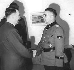 Theodor Eicke podczas odprawy z udziałem Adolfa Hitlera. Na zdjęciu widać dwóch, podających sobie dłonie, mężczyzn, z których Hitler ubrany jest w koszulę i marynarkę, a drugi mężczyzna, Eicke, w mundur formacji nazistowskiej z okresu Niemiec hitlerowskich. Za Hitlerem, niewidoczny, stoi trzeci mężczyzna.