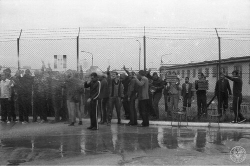 Ośrodek Odosobnienia w Kwidzynie, 14 sierpnia 1982 r. Grupa mężczyzn na placu podczas akcji protestacyjnej internowanych w tym ośrodku. Polewani są wodą prawdopodobnie z węża strażackiego. Część z nich ma uniesione ręce z palcami w geście „V”. Niektórzy trzymają drewniane taborety, prawdopodobnie się nimi zasłaniając. Fot. z zasobu IPN