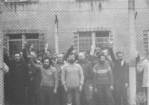 Akcja protestacyjna osób internowanych w Ośrodku Odosobnienia w Uhercach, 19 kwietnia 1982 r. Internowani stoją przy siatce ogrodzeniowej podczas akcji protestacyjnej; prawdopodobnie śpiewają. Część osób podnosi ręce, pokazując znak „V”, czyli znak zwycięstwa (victory). Na drugim planie budynek Zakładu Karnego w Uhercach. Fot. z zasobu IPN