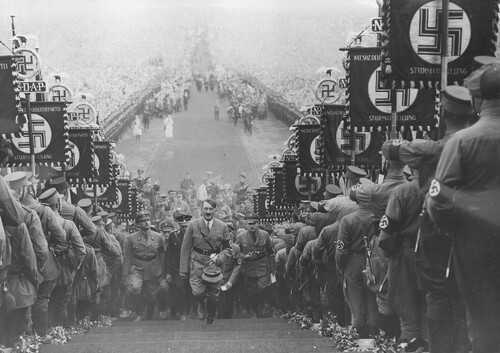 Uroczystości dożynkowe III Rzeszy, 30 września 1934 r. Adolf Hitler w otoczeniu oficerów wchodzi na Bückeberg pod Hameln (Dolna Saksonia) przez szpaler utworzony z partyjnych sztandarów. Na drugim planie widoczny wielotysięczny tłum Niemców. Ze zbiorów Narodowego Archiwum Cyfrowego
