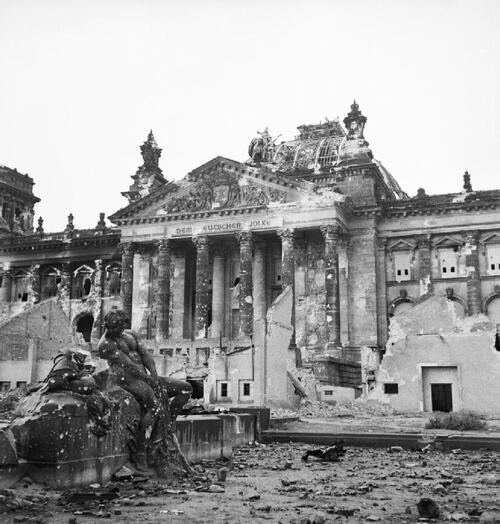 Widok fasady budynku wraz z fragmentem stojącego przed nim monumentu z sylwetką mężczyzny. Obiekt wraz z otoczeniem nosi ślady uszkodzeń i ostrzału. Wokół fragmenty gruzów.