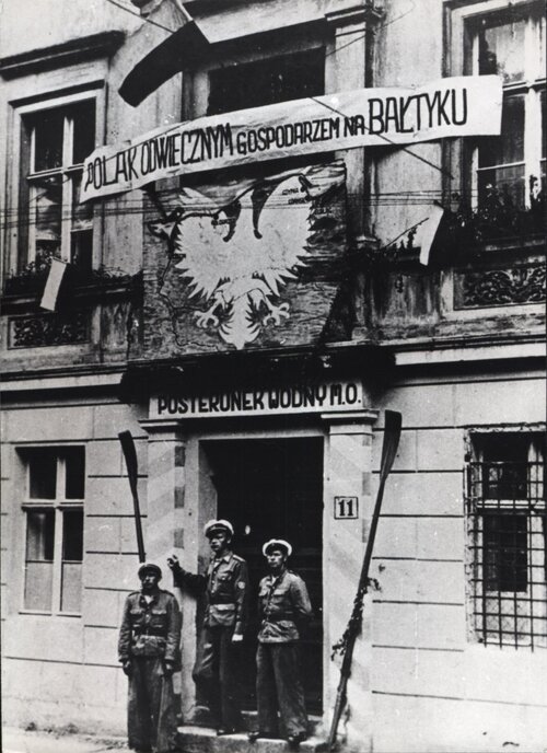 Trójka funkcjonariuszy przed wejściem do siedziby milicji. Nad wejściem okazała dekoracja z Orłem Białym i napisem: Polak odwiecznym gospodarzem na Bałtyku