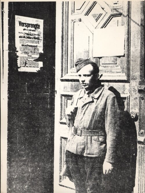 Mężczyzna w ubraniu przypominającym umundurowanie z bronią na ramieniu stoi przed drewnianymi drzwiami do budynku