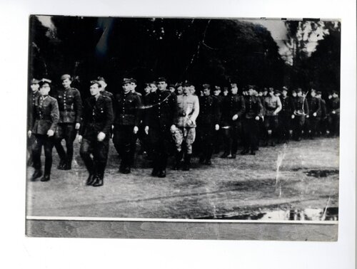 Żołnierze w szyku marszowym. W tle zarys dachu wiejskiego budynku