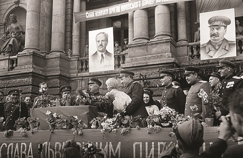 Grupa oficerów na trybunie ustawionej przed fasadą budynku przybranego propagandowymi portretami i transparentami z napisami w j. rosyjskim