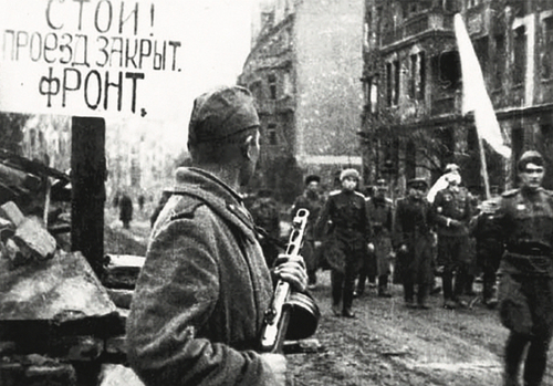 Na pierwszym planie mężczyzna stojący z karabinem, dalej grupa mężczyzn z białą flagą